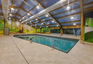 Piscina coperta in glamping, parete con murale foresta, scivolo verde, grandi finestre, luci moderne.