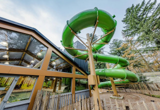 Grote groene waterglijbaan naast glazen gebouw in een vakantiepark met glamping-accommodaties.