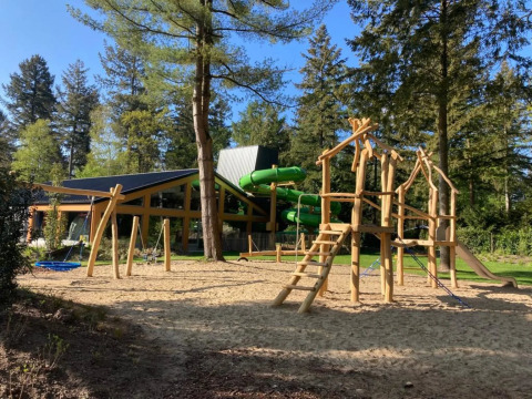 Parque infantil con estructuras de madera y tobogán en un camping glamping rodeado de bosque.