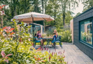 Familie nyder måltid udendørs under parasol ved luksuriøs glampinghytte i grøn feriepark.