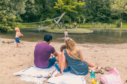 Familie nyder en solrig dag ved en sø i en feriepark med glamping, børn leger i vandet og sandet.