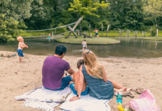 Familie nyder en solrig dag ved en sø i en feriepark med glamping, børn leger i vandet og sandet.