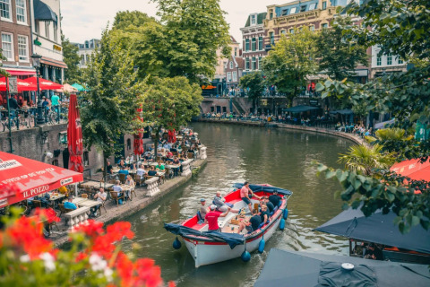 People dining at lively canal-side cafes as a small boat cruises through a scenic European city center.