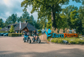 Entrance to EuroParcs holiday park with two people riding electric scooters in front of the main building.