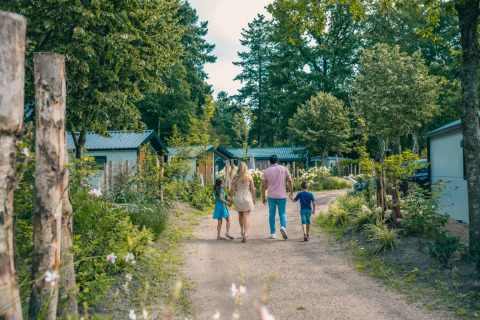 Familie går ad en grussti i et feriepark med glampinghytter omgivet af grønne træer og natur.
