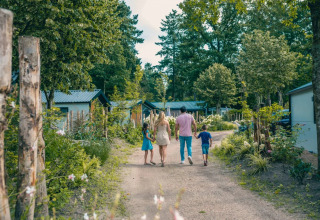 Familie går ad en grussti i et feriepark med glampinghytter omgivet af grønne træer og natur.