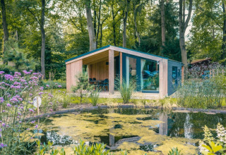 Et moderne glampinghus i træ med overdækket terrasse, omgivet af skov og dam i en feriepark.