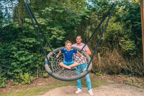 Niño sentado en un gran columpio, empujado por un adulto, rodeados de naturaleza en un parque de glamping.