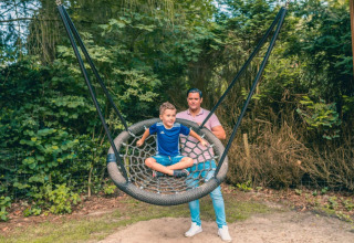Kind auf einer großen Schaukel mit einem Erwachsenen dahinter, umgeben von Bäumen in einem Glamping-Park.