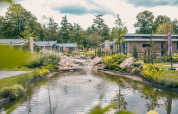Idyllisk feriested med glamping, små hytter, dam med springvand og grønne omgivelser under overskyet himmel.