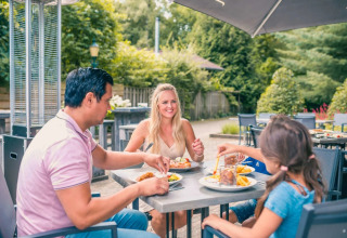En familie nyder et måltid udendørs på et feriepark med glamping, omgivet af grønne træer og natur.