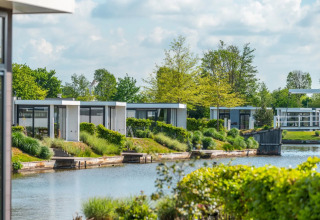 Moderne glamping-hytter ved vandet i en feriepark, omgivet af grøn beplantning og træer.