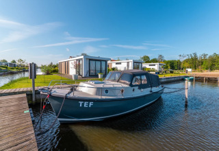Moderne glamping-hytter ved en sø med privat bådebro og en fortøjet båd under en klar blå himmel.