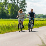 Twee mensen fietsen op een landweg bij een vakantiepark met luxe glamping-accommodaties in het groen.
