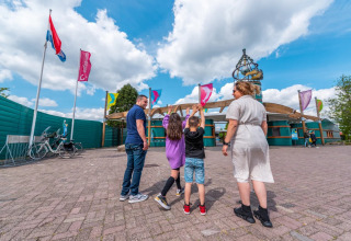 En familie ankommer til et feriepark med glamping, omgivet af flag og en klar, blå himmel.