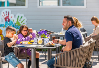 Familie nyder udendørs frokost ved ferieparkens glamping-område med børn, sol og afslappet stemning.