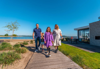En familie går ved en sø i en feriepark med glamping-indkvartering, solskin og blå himmel.