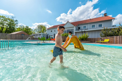 Bambini giocano in una piscina bassa con scivolo presso un glamping park, con cottage moderni sullo sfondo.