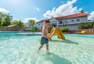 Børn leger i lav swimmingpool med rutsjebane ved glamping feriepark med moderne hytter i baggrunden.