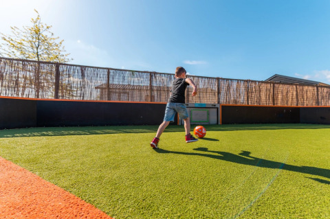 Jongen speelt voetbal op kunstgrasveld in vakantiepark met glamping accommodaties onder blauwe lucht.