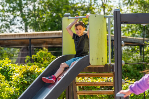 Niño deslizándose por un tobogán en un parque de vacaciones con glamping, rodeado de plantas verdes.