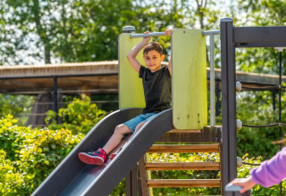Garçon glissant sur un toboggan de terrain de jeu dans un parc de vacances avec glamping, entouré de verdure.