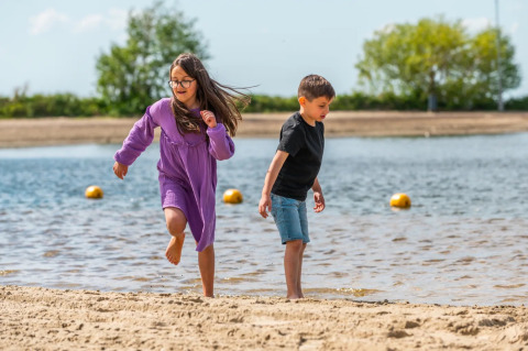 Dos niños juegan en la orilla de una playa en un parque vacacional con opciones de glamping bajo el sol.