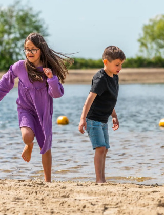Dos niños juegan en la orilla de una playa en un parque vacacional con opciones de glamping bajo el sol.