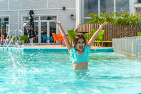 Fröhliches Mädchen spielt im Pool eines Ferienparks mit Glamping, bunte Stühle im Hintergrund.