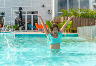 Niña feliz jugando en la piscina de un parque vacacional con glamping, sillas coloridas atrás.