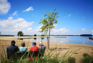 Vier mensen zitten op een bankje aan het zandstrand en kijken uit over het meer en de blauwe lucht.
