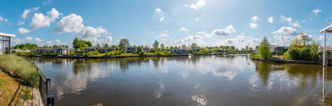 Vista panorámica de un parque vacacional con alojamientos glamping, cabañas modernas y lago tranquilo bajo el cielo azul.