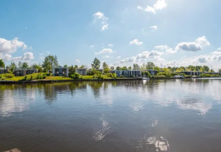 Panoramic view of a holiday park with glamping accommodations, modern cabins, and calm lake under blue sky.