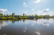Panoramic view of a holiday park with glamping accommodations, modern cabins, and calm lake under blue sky.