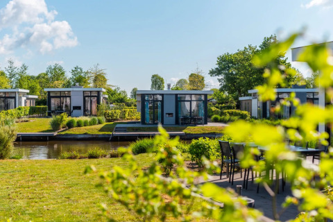 Moderne glampinghuisjes aan het water in een vakantiepark met groen en zonlicht op de achtergrond.