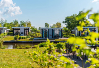 Moderne glampinghuisjes aan het water in een vakantiepark met groen en zonlicht op de achtergrond.