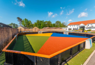 Terrain sportif coloré dans un parc de vacances avec hébergements glamping sous ciel bleu et soleil.