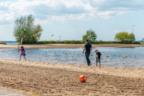Gezin aan het strand van een glamping vakantiepark, kinderen spelen nabij het water met een bal op het zand.