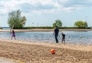 Family enjoys a beach at a glamping holiday park, with children playing near the water and a ball in the sand.