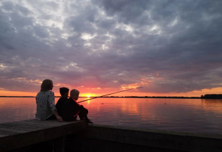 En familie sidder på en badebro ved søen og fisker sammen, mens solen går ned over vandet i en feriepark.