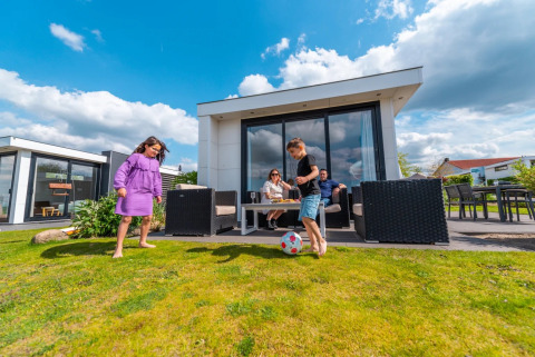 Niños juegan al fútbol sobre césped frente a una cabaña glamping moderna, padres descansan en la terraza.