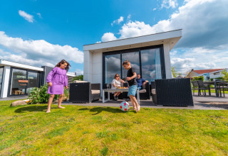 Children play soccer on the grass outside a modern glamping cabin while parents relax on the patio furniture.