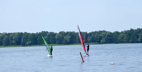 Due persone fanno windsurf su un lago calmo vicino a una foresta, in un parco vacanze glamping.