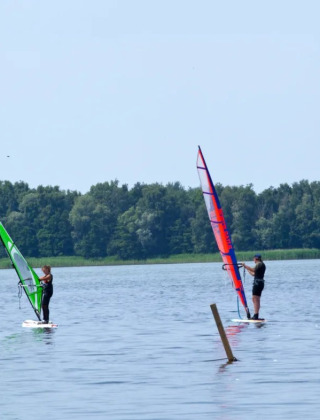 Dos personas haciendo windsurf en un lago tranquilo cerca de un bosque en un parque de vacaciones glamping.