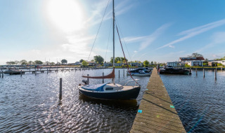 Día soleado en parque vacacional con velero amarrado en muelle de madera y alojamientos glamping al fondo.