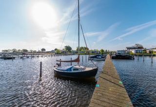 Sunny day at a holiday park marina with a sailboat docked at a wooden pier and glamping cabins behind.
