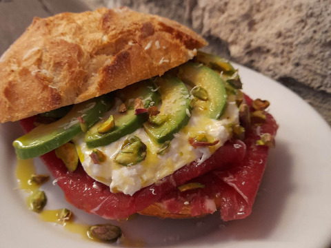 Sándwich gourmet con aguacate, burrata, pistachos y atún crudo en pan crujiente, servido en plato blanco.