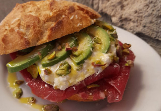 Gourmet-sandwich med avocado, burrata, pistacienødder og rødt kød på sprødt brød, serveret på en tallerken.
