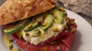 Sándwich gourmet con aguacate, burrata, pistachos y atún crudo en pan crujiente, servido en plato blanco.