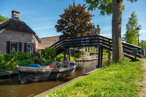 Personas navegan en barco por un canal junto a una casa y un puente en un parque de vacaciones glamping.